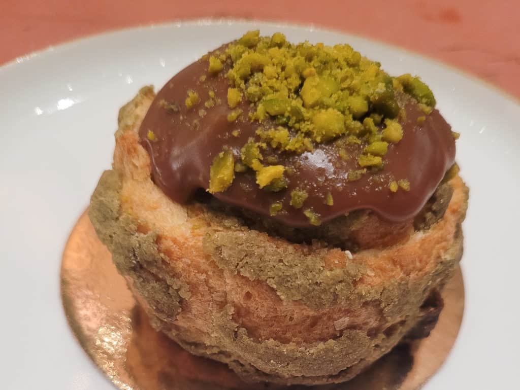 Pistachio cream puff topped with chocolate and pieces of pistachio on a white plate