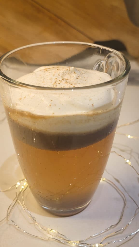 A clear mug with hot buttered rum topped with whipped cream surrounded by string lights.