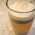 A closeup of a clear mug with hot buttered rum topped with whipped cream surrounded by string lights.