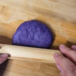 Two hands holding a small rolling pin flattening ube pandesal dough