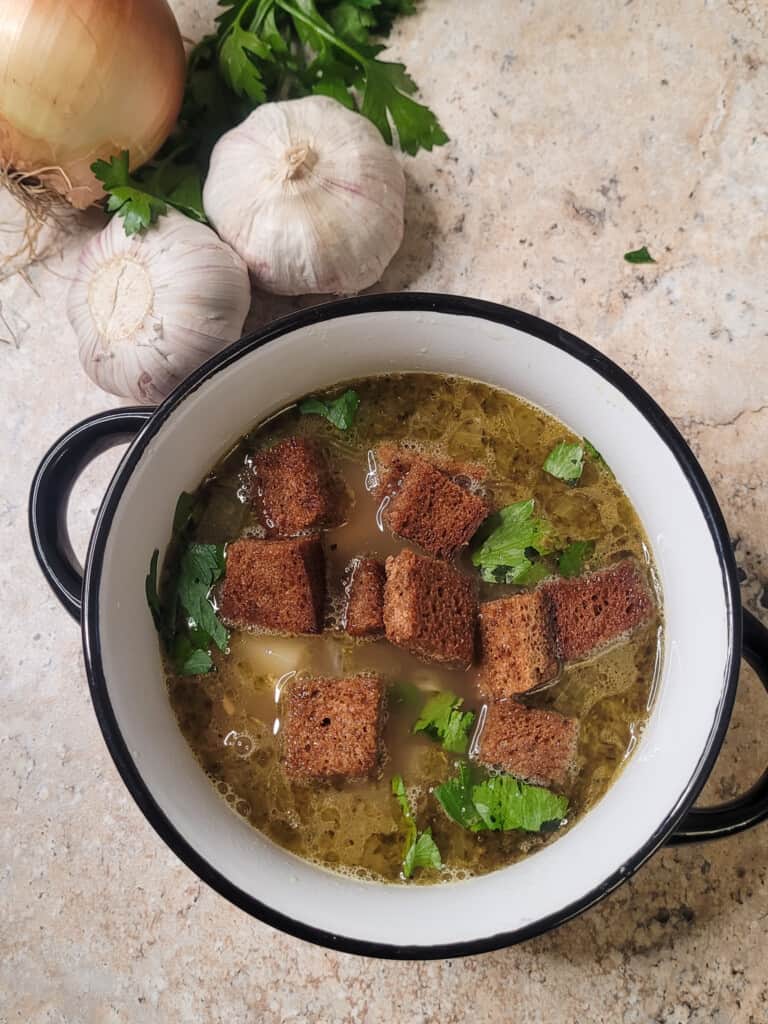 Garlic comfort soup in a bowl next to an onion and bulbs of garlic