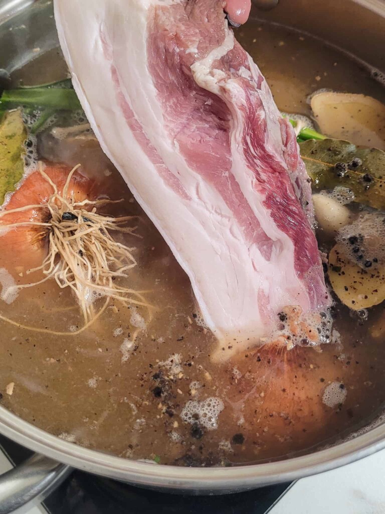 A slice of pork belly lowered into a pot of boiled water