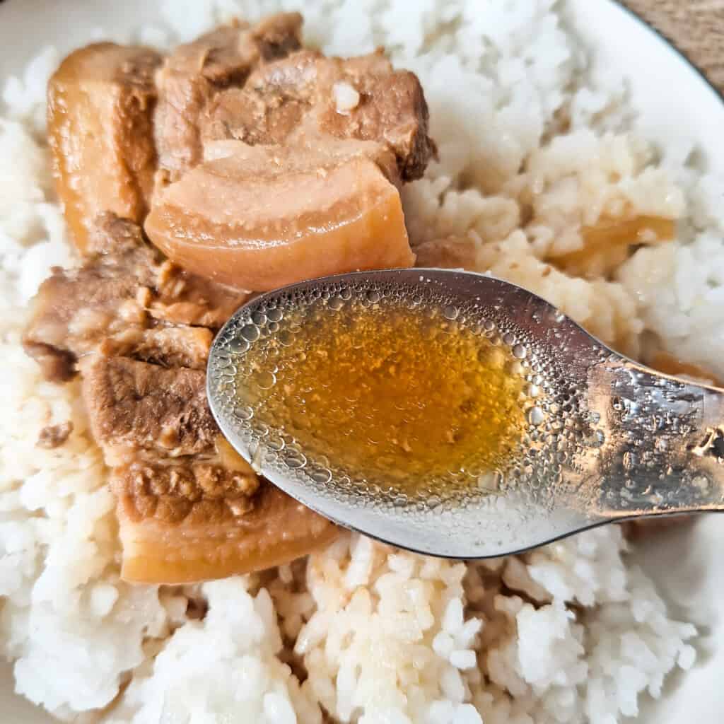 Filipino adobo soup in a spoon in front of a plate of adobo with rice.