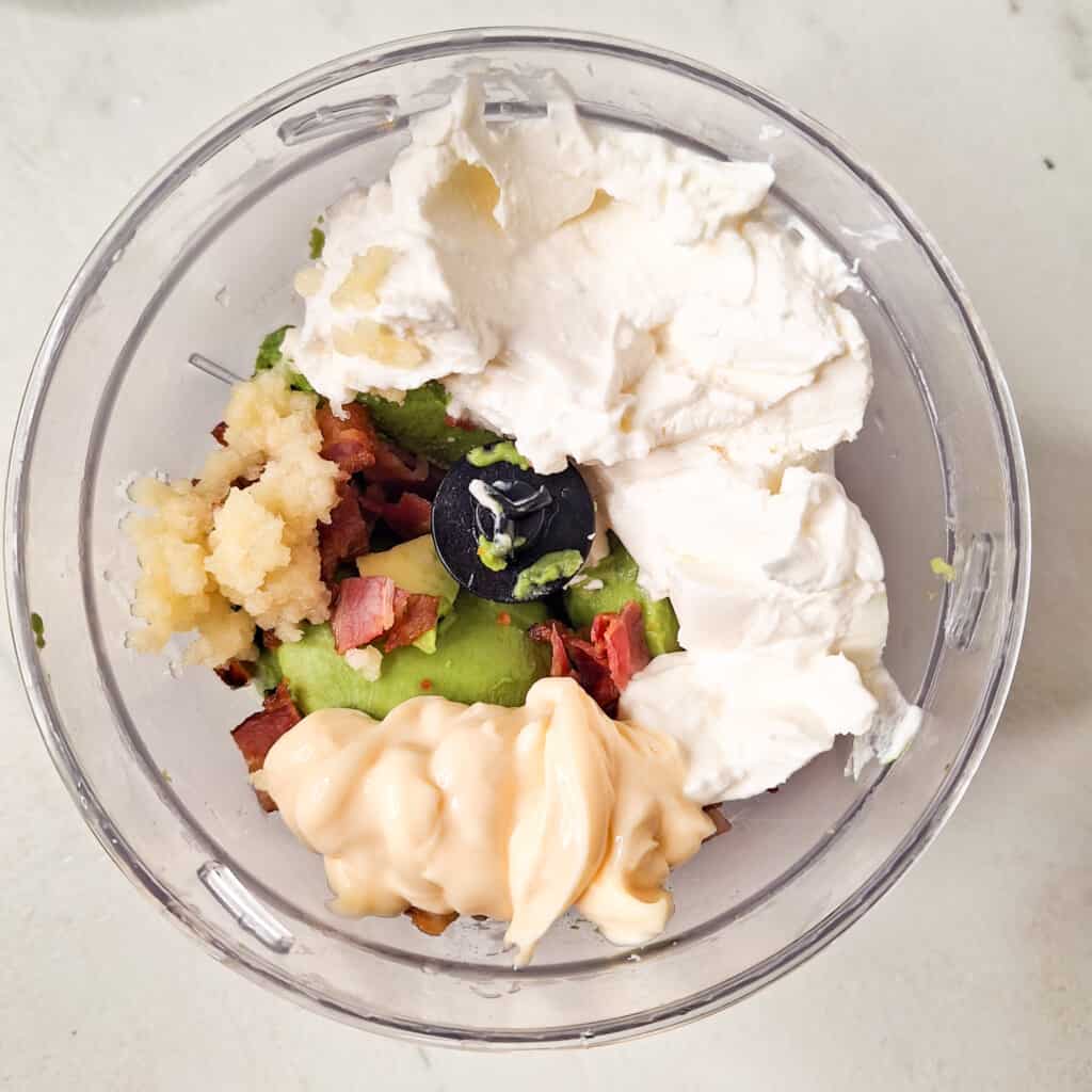 Overhead view of ingredients in a food processor that includes kewpie mayo, avocado, garlic, cream cheese, and bacon