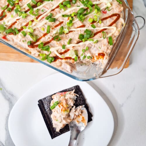 A spoon with a nor sheet and some fish and rice next to a tray of sushi bake