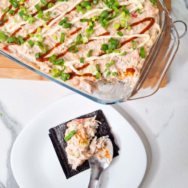 A spoon with a nor sheet and some fish and rice next to a tray of sushi bake